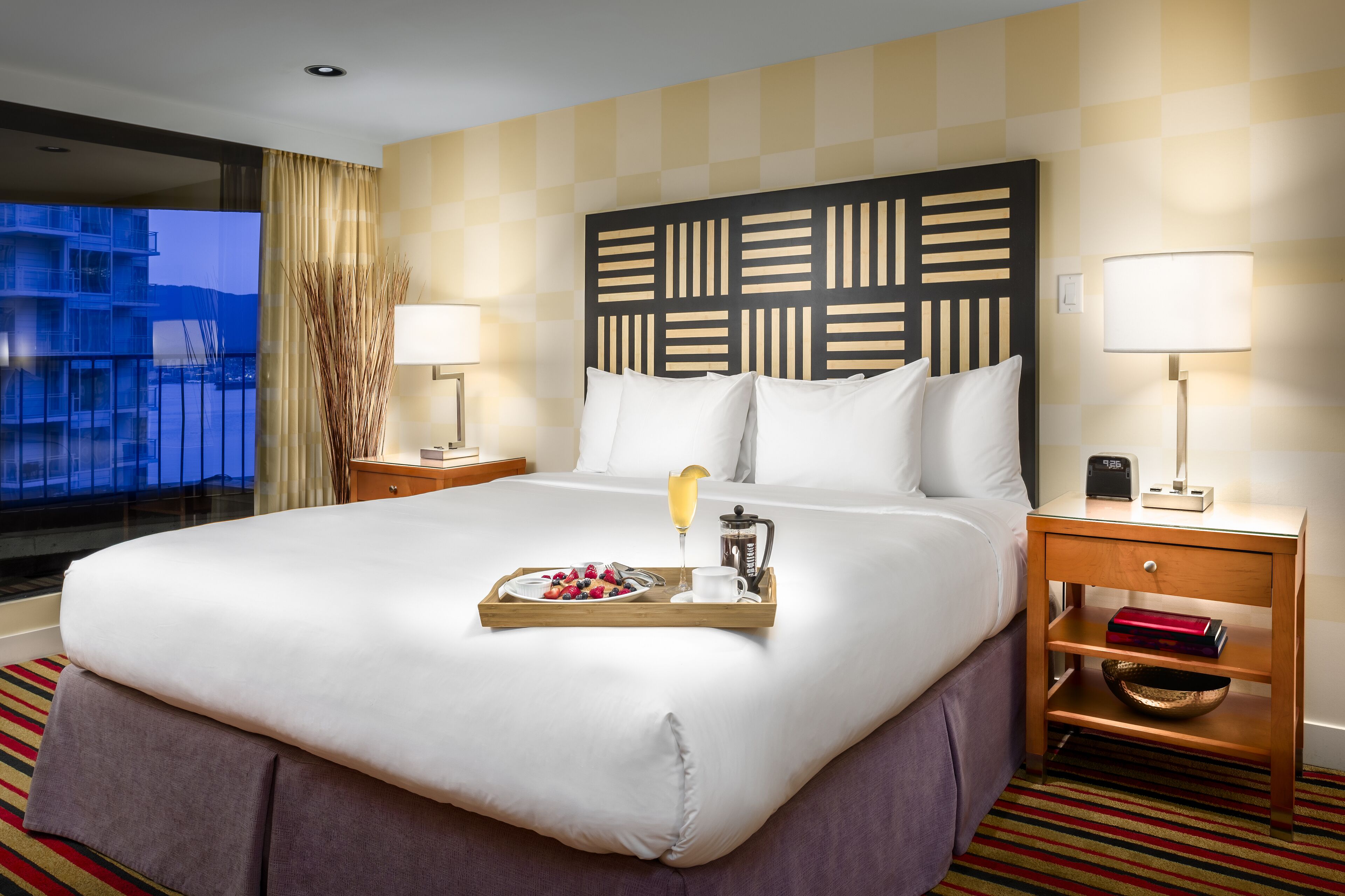 A hotel room with a large bed, a wooden headboard, two bedside tables with lamps, a tray with breakfast items, and a view of buildings through a window.
