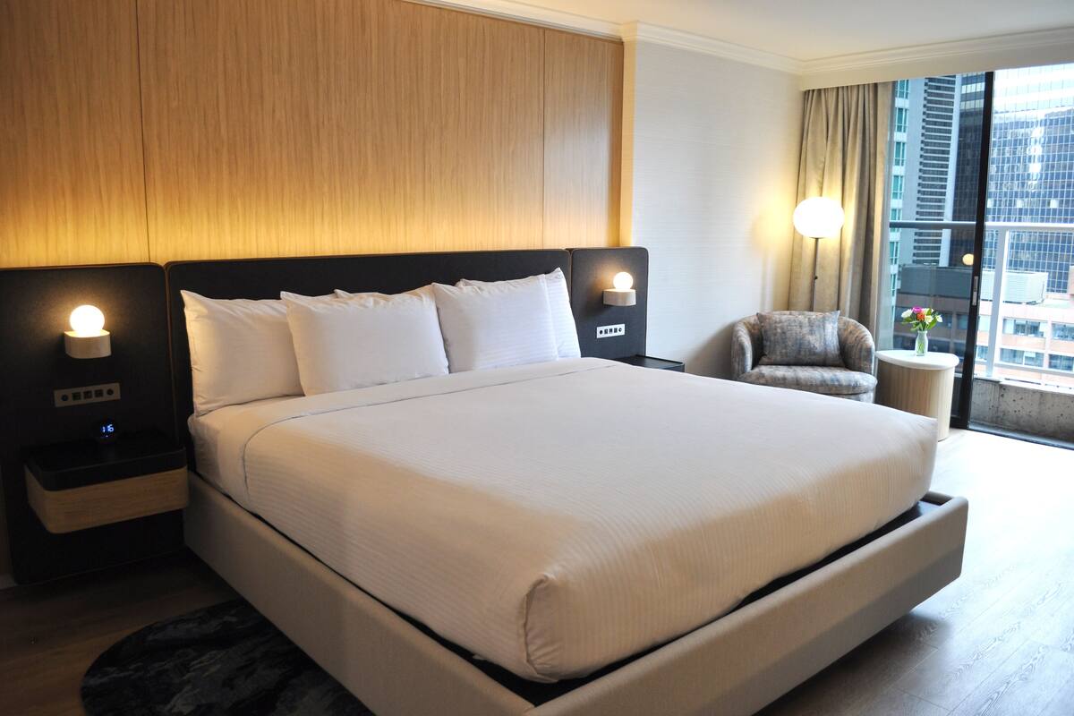 A hotel room with a large bed, bedside lamps, a chair, and a window with a city view.