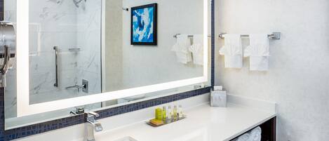 Un baño moderno con un espejo grande, un lavamanos y una ducha. Hay toallas y artículos de aseo en el mostrador.
