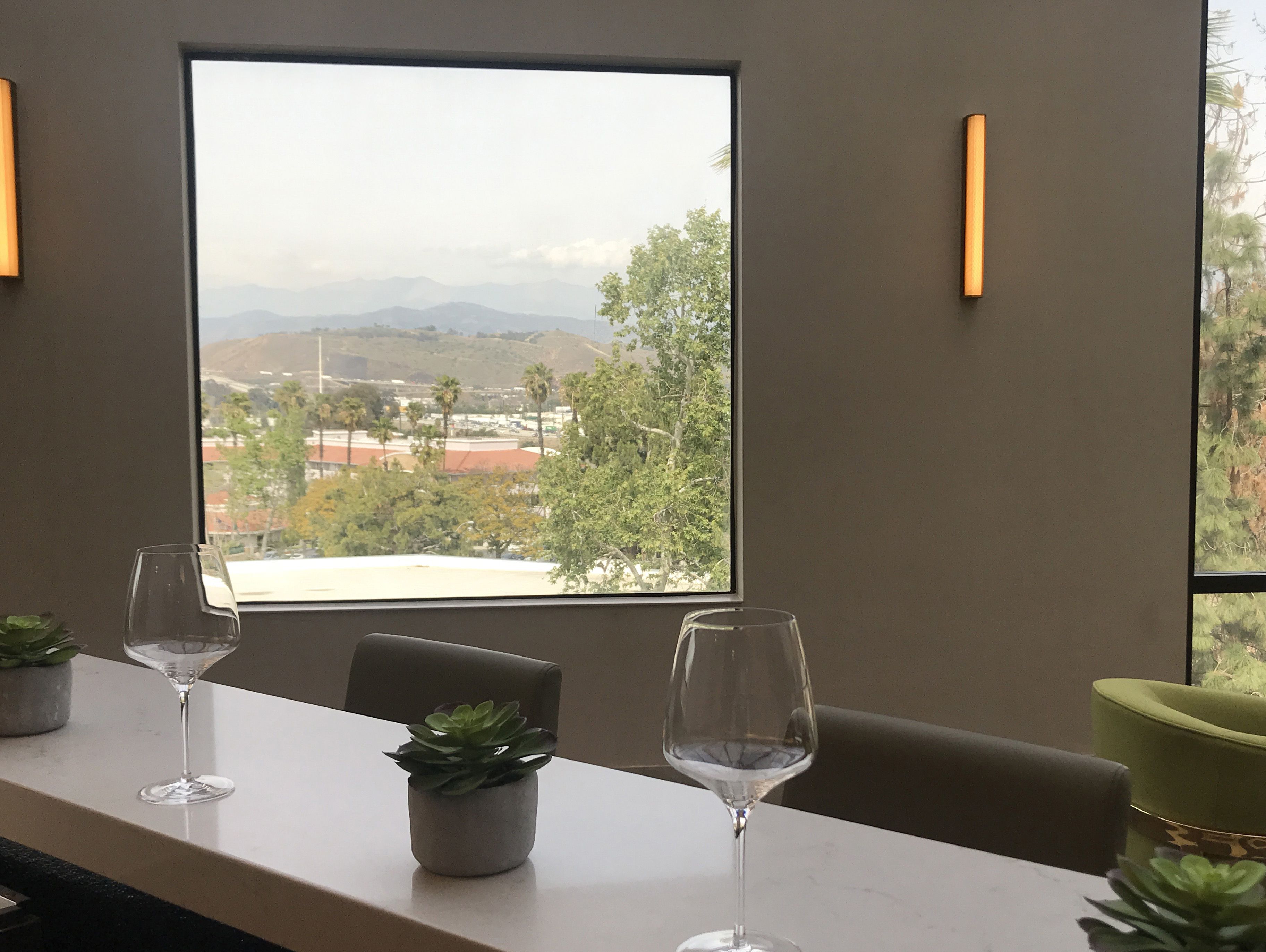 Quarto com uma janela ampla que oferece vista para a paisagem urbana e montanhas. Há duas taças de vinho vazias sobre uma mesa, um vaso pequeno com planta e uma cadeira verde.