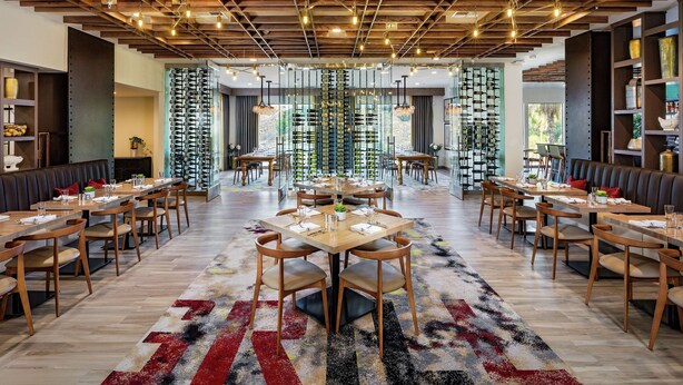 A spacious restaurant with multiple dining tables and chairs, wooden ceiling beams, and a colorful rug. - DoubleTree by Hilton Pomona (Pomona)