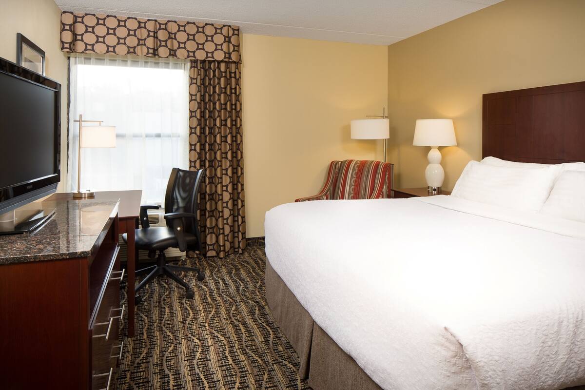 A hotel room with a bed, a desk with a television, a chair, two lamps, and a patterned carpet.