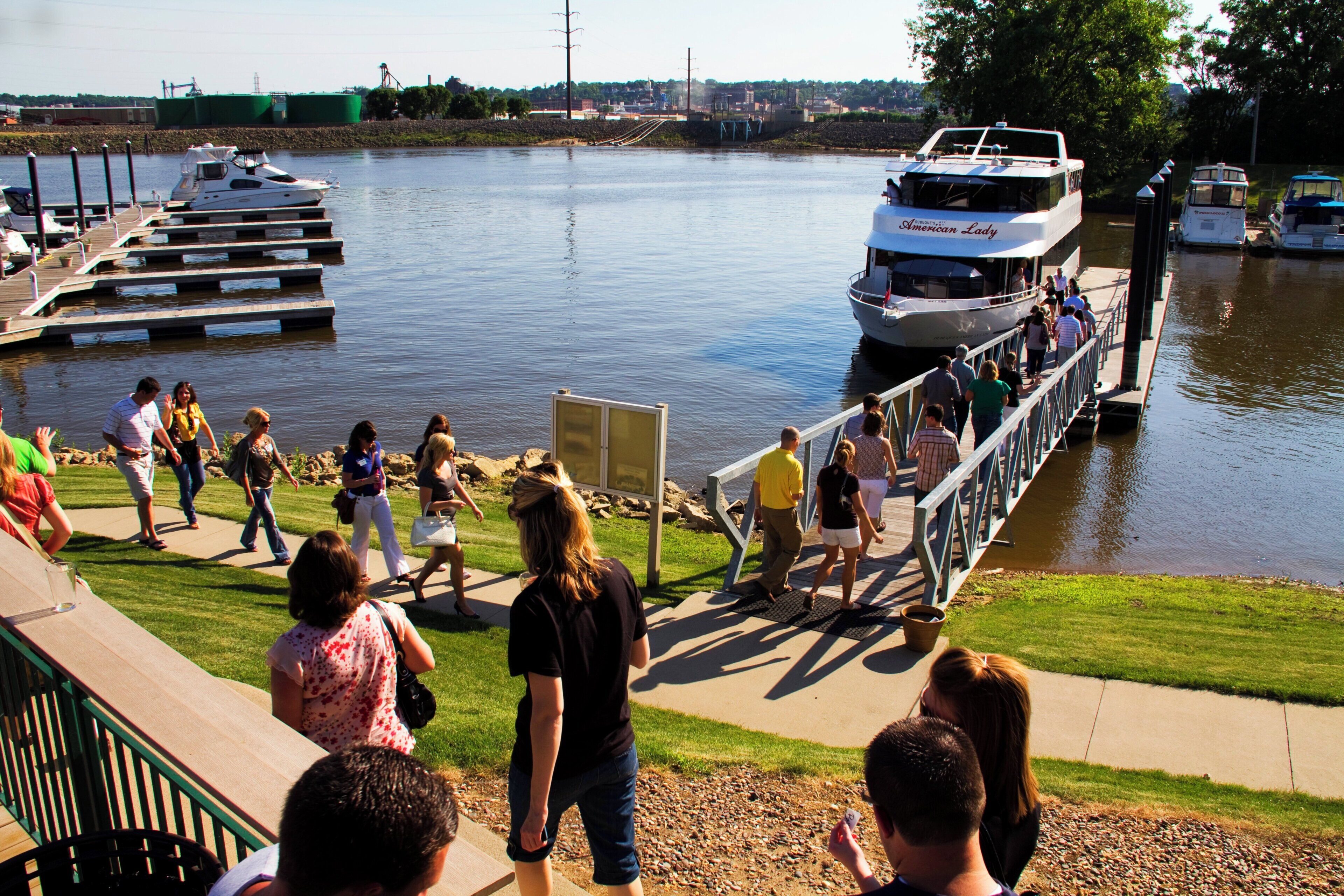 A waterfront promenade with people walking, a dock with boats, and a clear view of the water.