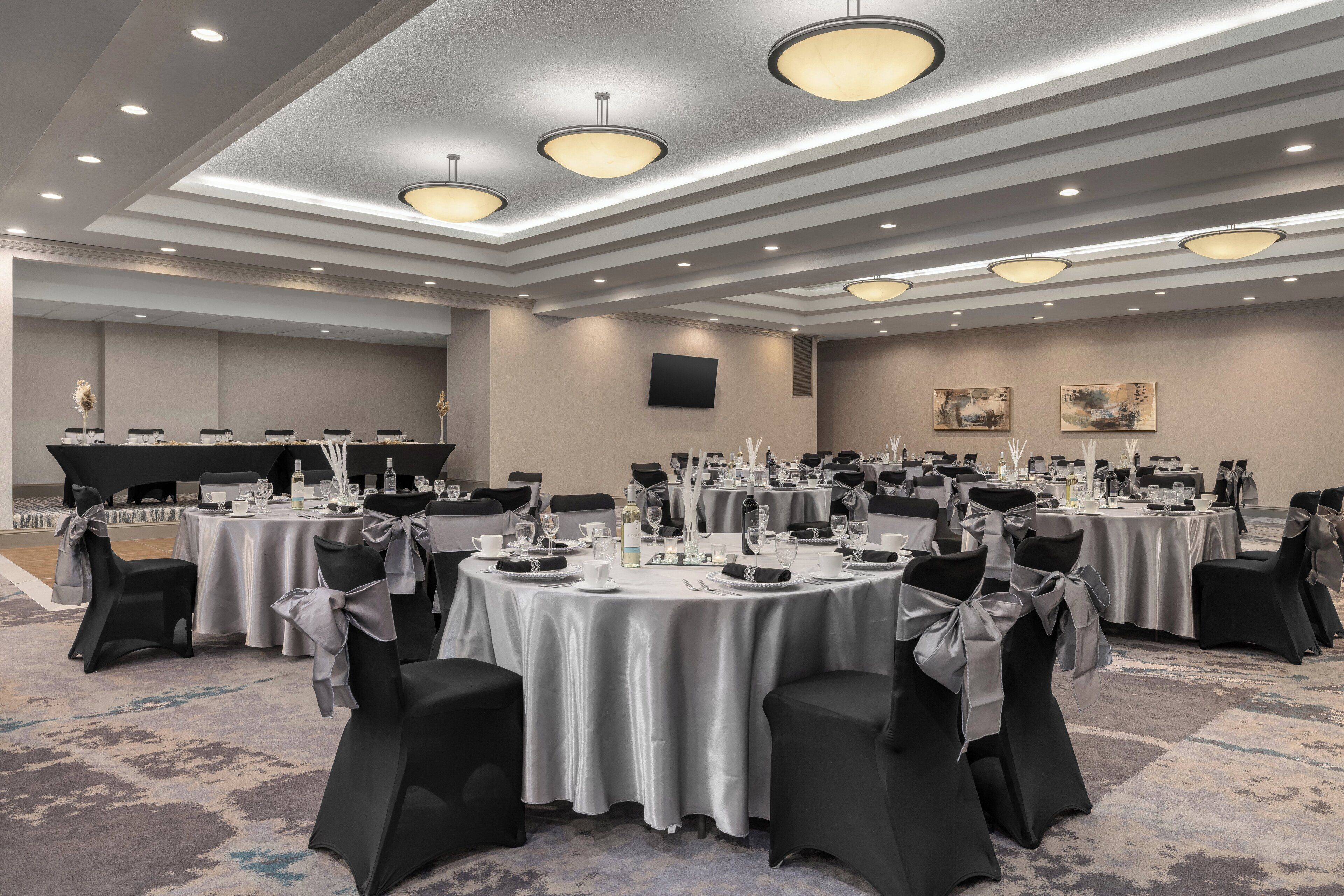 Une salle de banquet avec des tables rondes, des chaises noires ornées de nœuds argentés et un plafond au design moderne.