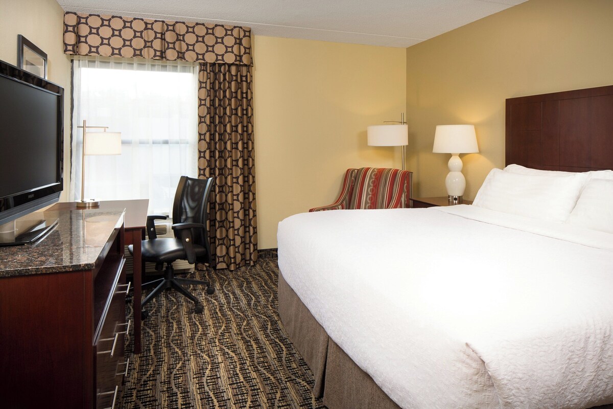 A hotel room with a bed, a desk with a television, a chair, two lamps, and a patterned carpet.