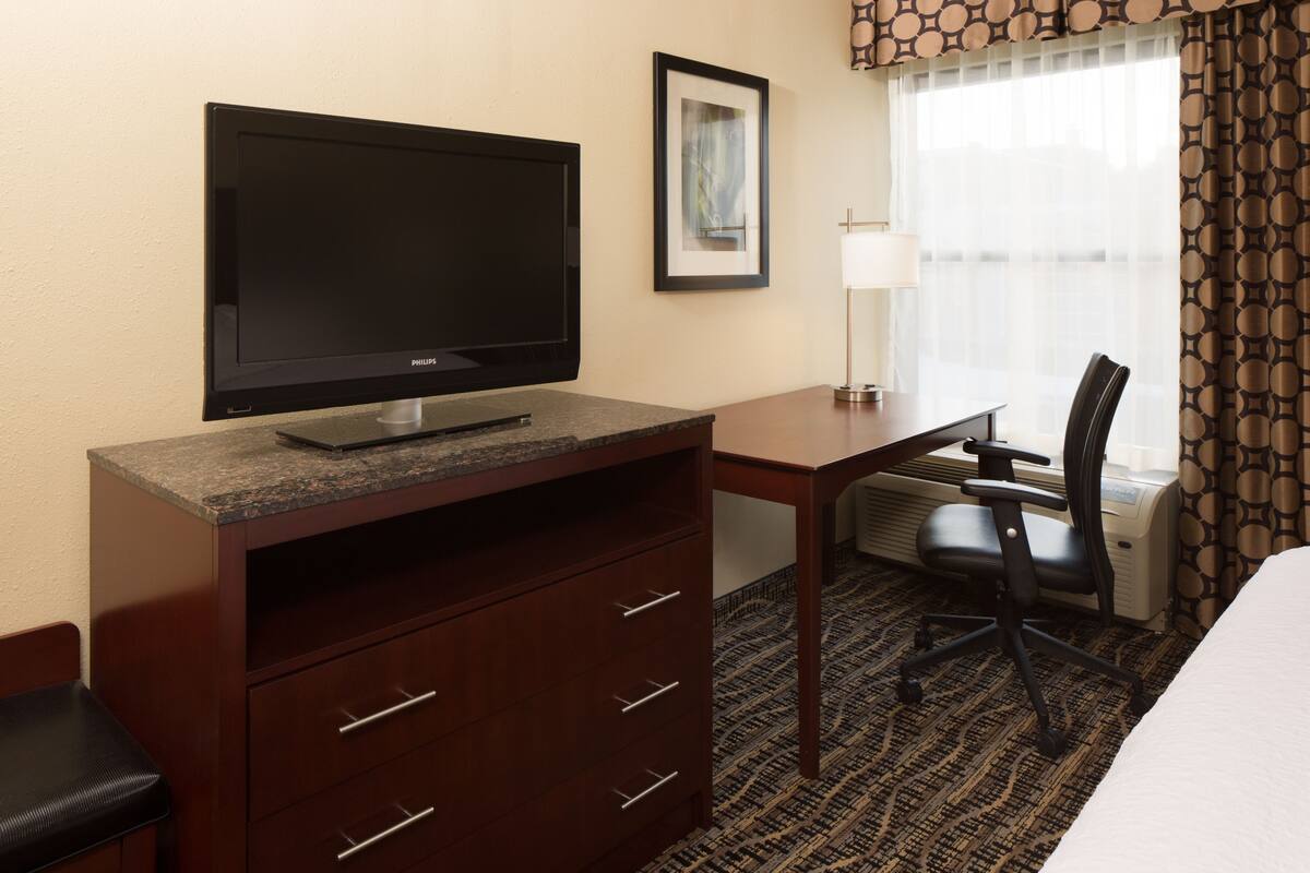 A hotel room with a flat-screen TV, a desk with a chair, a bed, a framed picture on the wall, and a window with curtains.