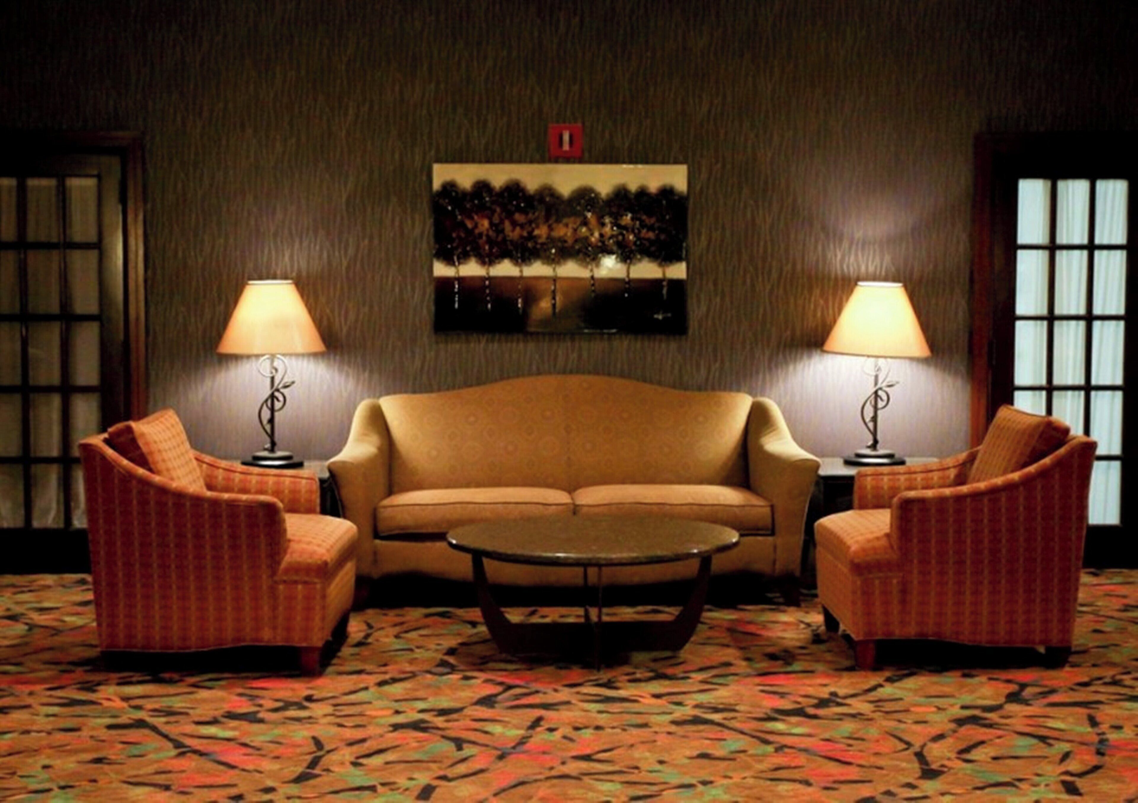 A room with a sofa, two armchairs, a coffee table, and two lamps. There is a painting on the wall and a patterned carpet.