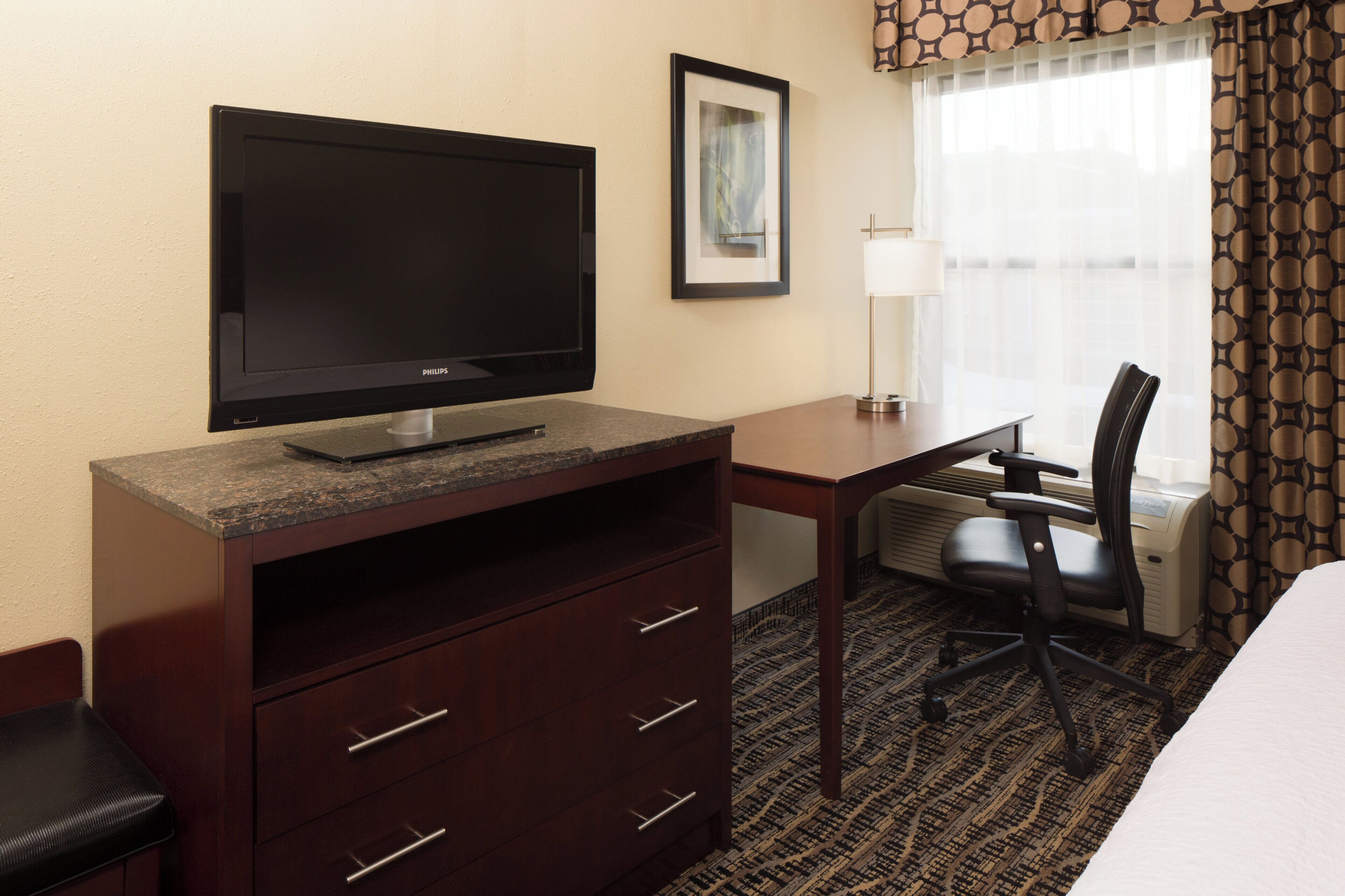 A hotel room with a flat-screen TV on a wooden desk, a chair, a bed, and a window with curtains.