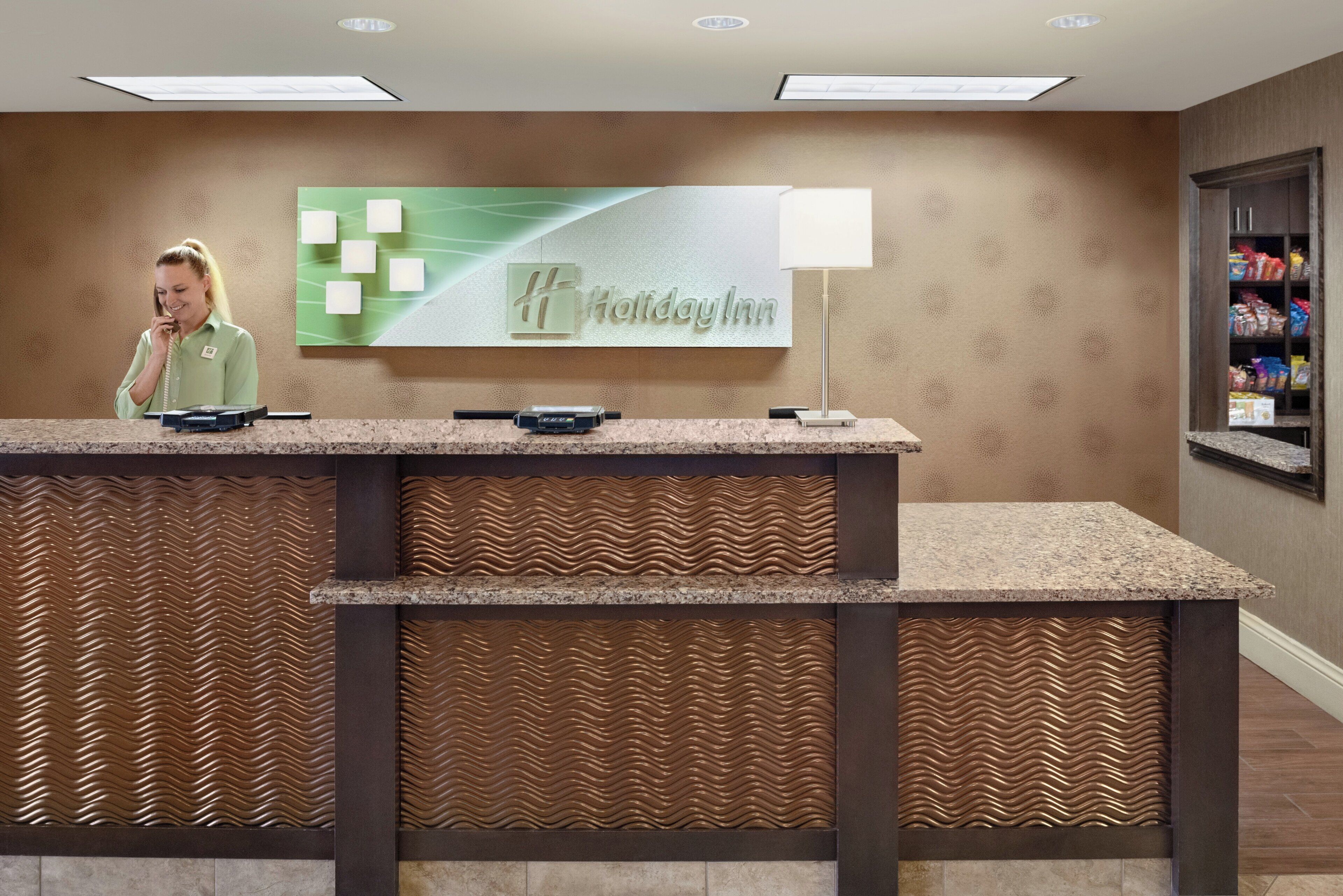 Mostrador de recepción con placa de Holiday Inn, una recepcionista y una barra de snacks con productos envasados.