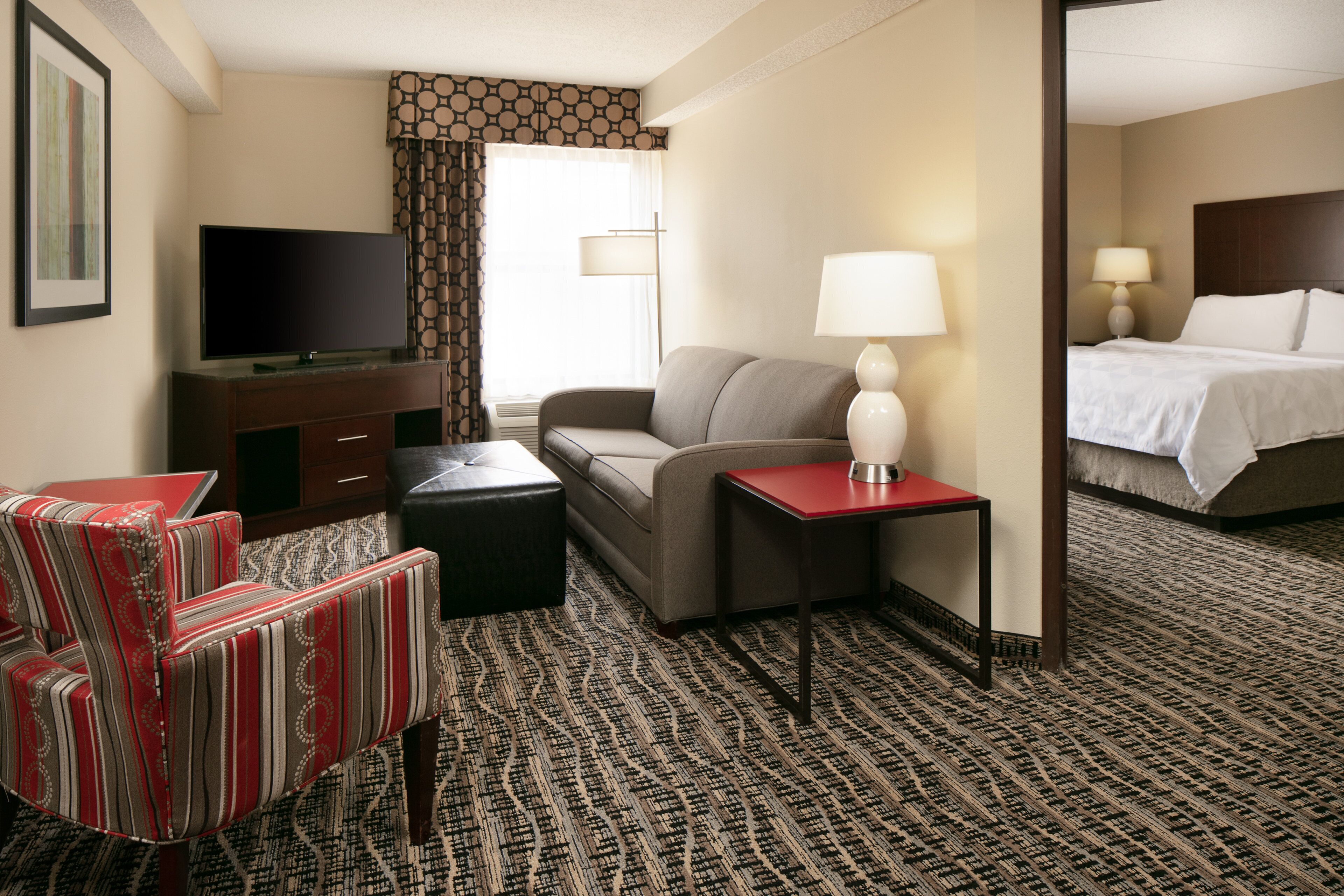 A hotel room with a sofa, armchair, bedside table, lamp, television, and a bed.