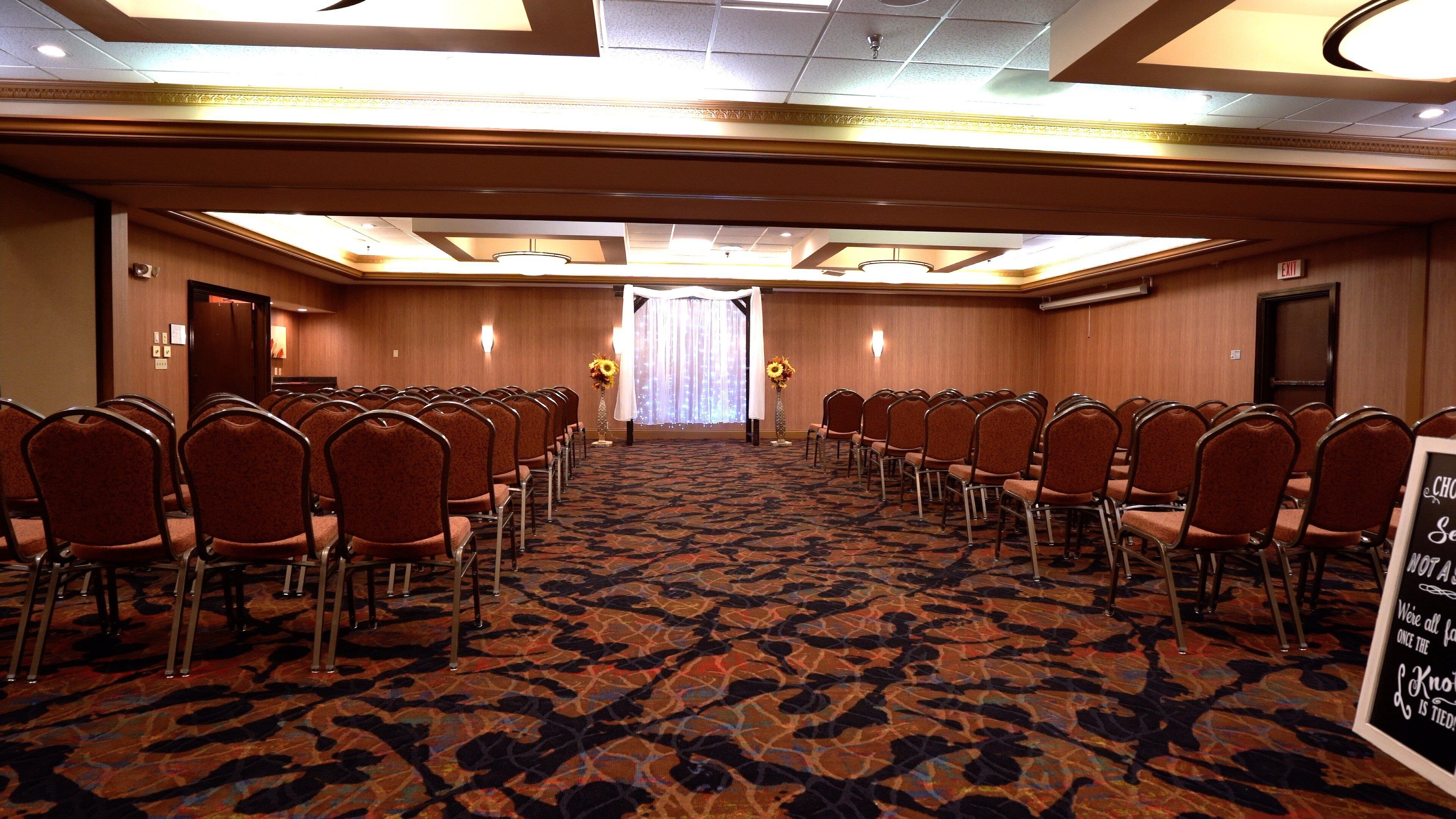 Un salón de conferencias con filas de sillas, piso alfombrado y un amplio ventanal con cortinas.