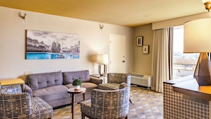 Ein Hotelzimmer mit einer Couch, zwei gemusterten Sesseln, einem kleinen Tisch mit einer Pflanze, einer Lampe, einem Gemälde von einer Berglandschaft, einer Tür und einem Fenster mit Vorhängen.