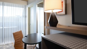 Ein Hotelzimmer mit Bett, Tisch, Stuhl, Lampe und einem Fenster mit durchsichtigen Vorhängen.