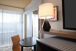 A hotel room with a bed, a table, a chair, a lamp, and a window with sheer curtains.