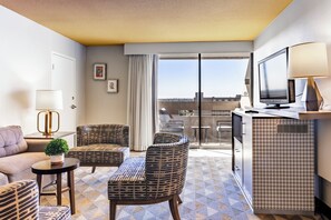 A hotel room with a sofa, patterned chairs, a small table with a plant, a TV, and a balcony with a view.