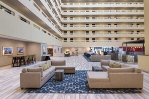A spacious hotel lobby with multiple seating areas, a central staircase, and a view of the upper floors.