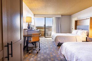 A hotel room with two beds, a desk with a chair, a lamp, and a view through a glass door.