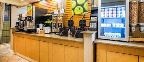 Un mostrador de autoservicio con una máquina de café, una máquina para hacer gofres y un refrigerador con bebidas y aperitivos.