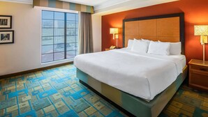 A hotel room with a large bed, a window with curtains, a patterned carpet, and a nightstand with a lamp.