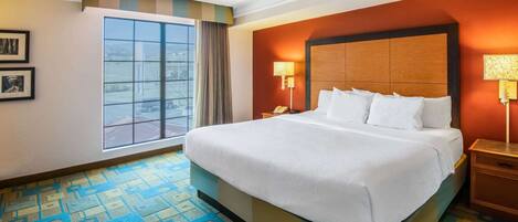 A hotel room with a large bed, a window with curtains, a patterned carpet, and a nightstand with a lamp.