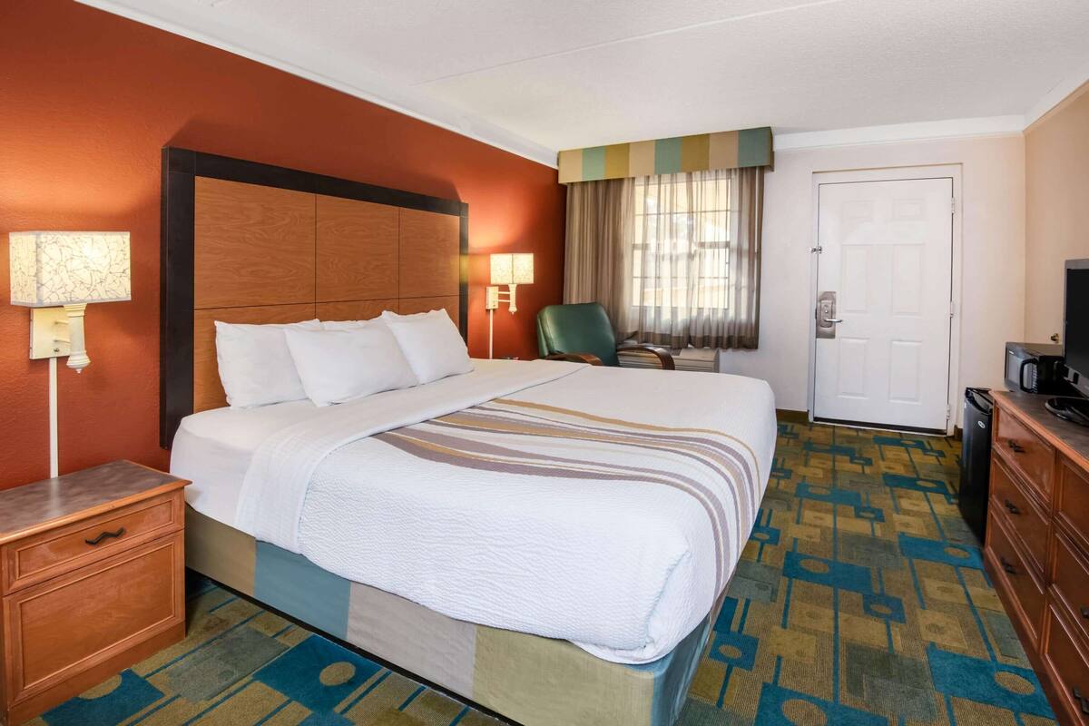 A hotel room with a large bed, a wooden headboard, a nightstand, a desk, a chair, a television, and a door.