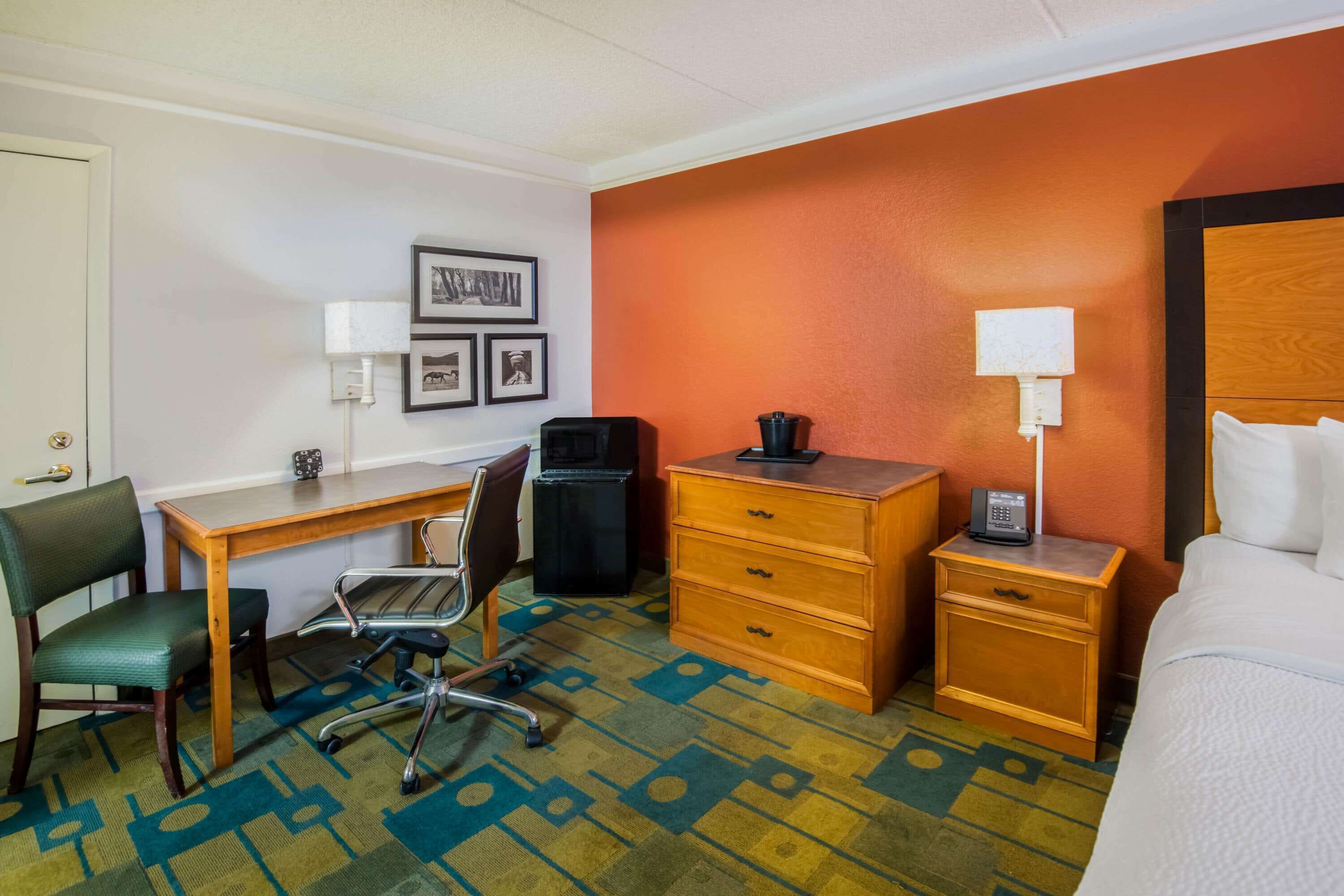 A hotel room with a bed, a desk with a chair, a dresser, a nightstand with a phone, and a wall-mounted lamp.