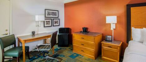 A hotel room with a bed, a desk with a chair, a dresser, a nightstand with a phone, and a wall-mounted lamp.