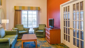 A room with two green armchairs, a wooden coffee table, a television on a wooden cabinet, and a large window with curtains.