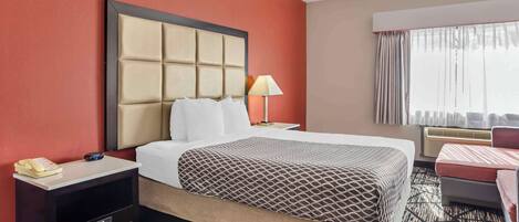 A hotel room with a large bed, a bedside table with a lamp, a telephone, and a small desk with drawers.