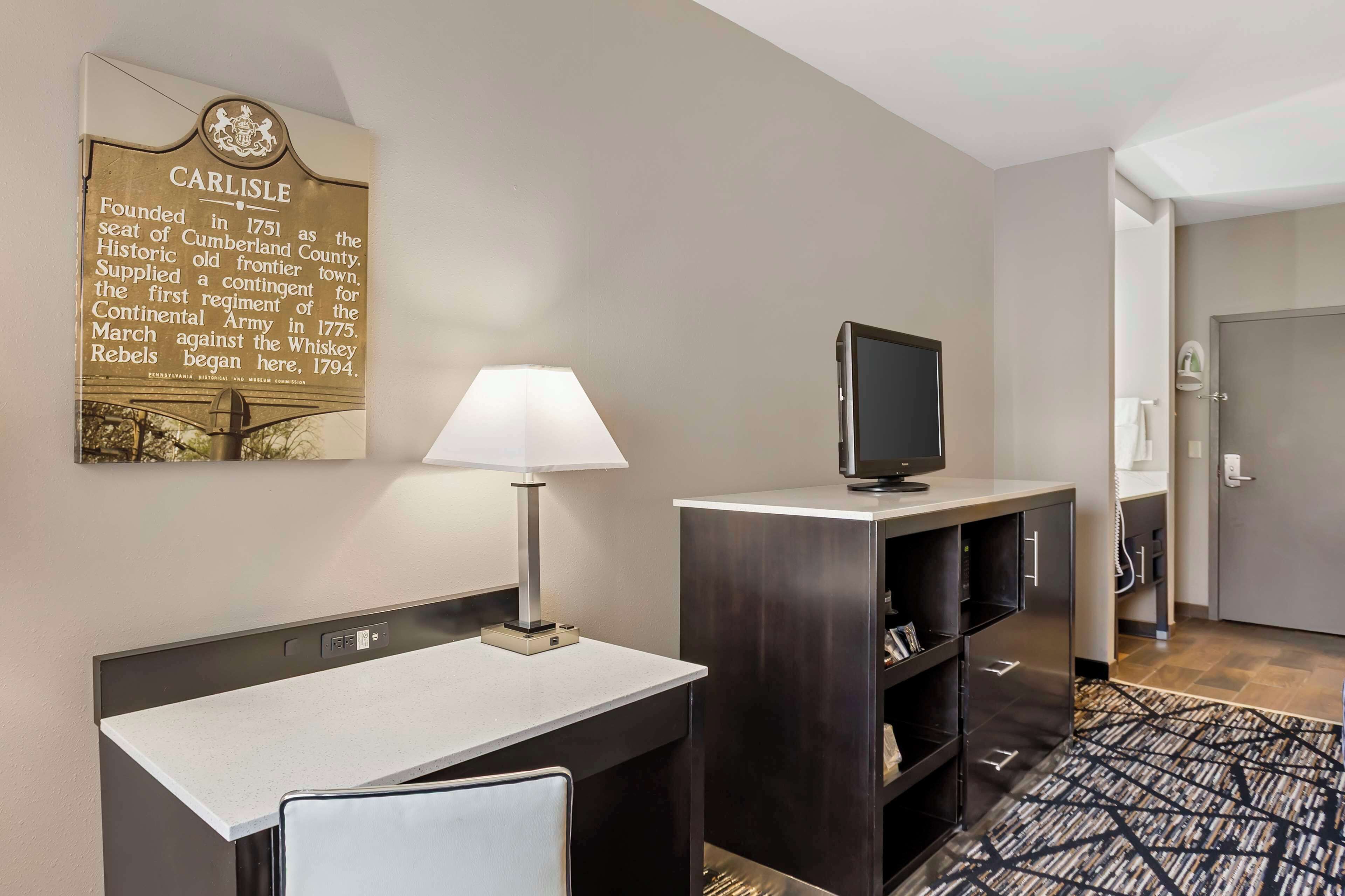 A hotel room with a desk, chair, lamp, television, and a wall plaque.