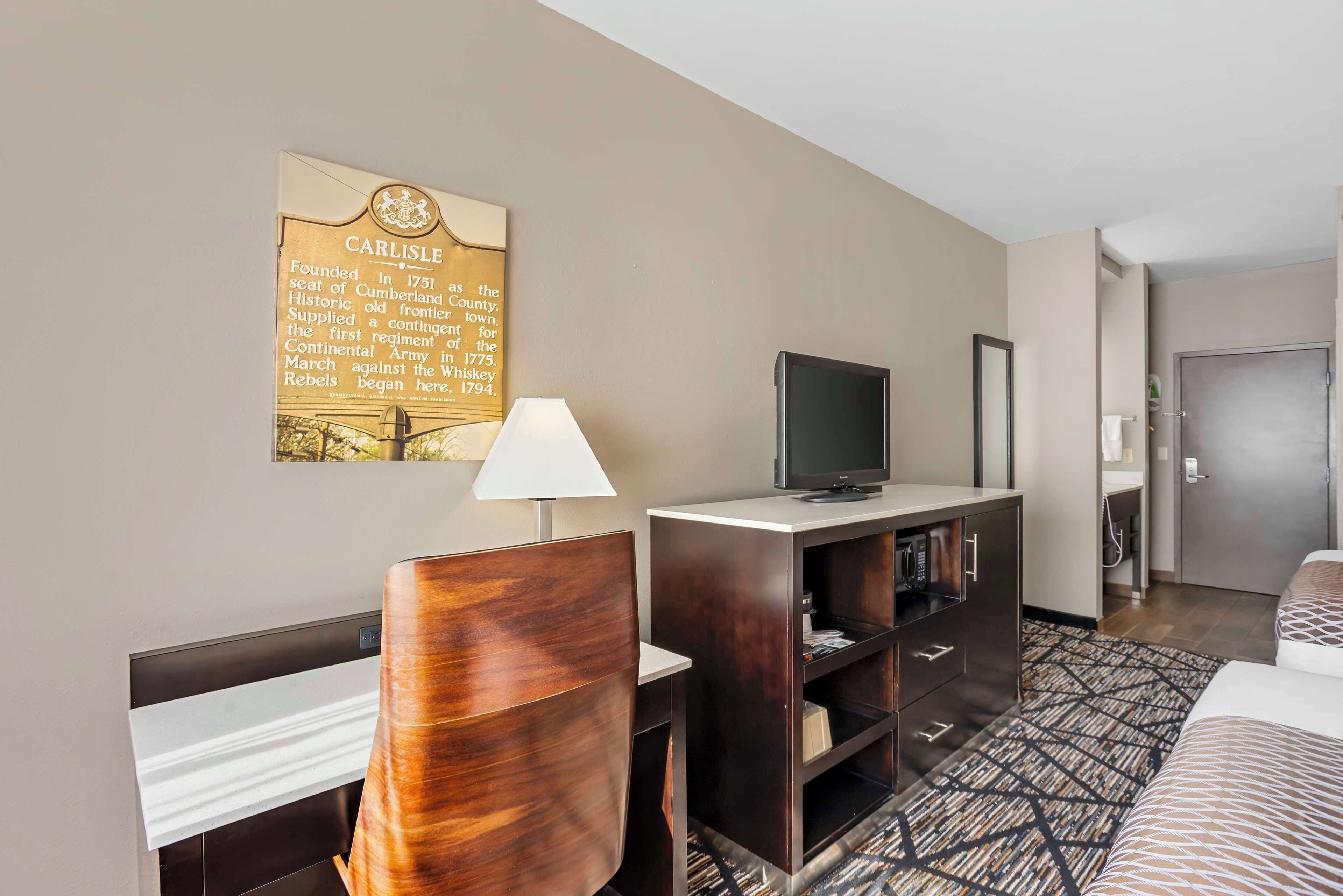 A hotel room with a wooden chair, a desk, a television, a bookshelf, and a framed historical plaque on the wall.