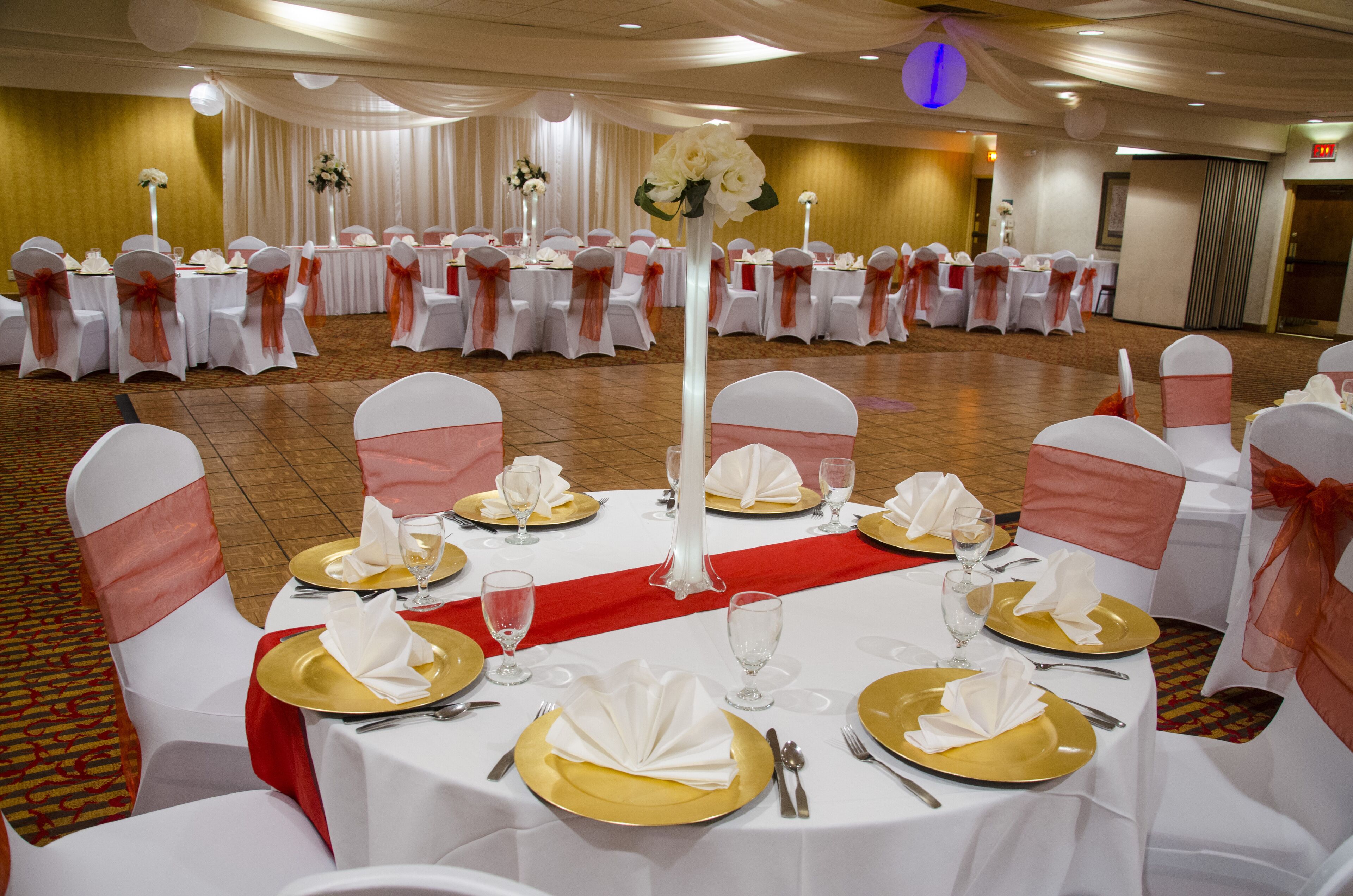 Une salle de banquet avec des tables rondes dressées pour un événement formel, agrémentée de nappes blanches, d’assiettes décoratives dorées et de rubans rouges sur les chaises.