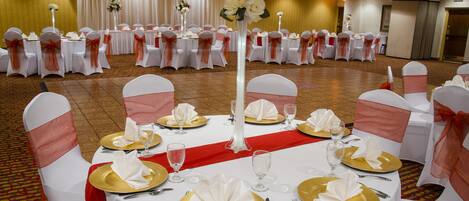 Une salle de banquet avec des tables rondes dressées pour un événement formel, agrémentée de nappes blanches, d’assiettes décoratives dorées et de rubans rouges sur les chaises.