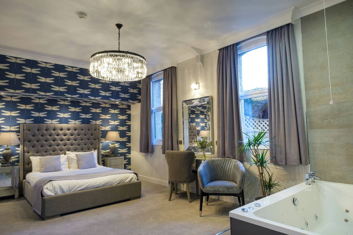 A bedroom with a tufted headboard bed, a sitting area with a chair and table, a large mirror, and a bathtub.