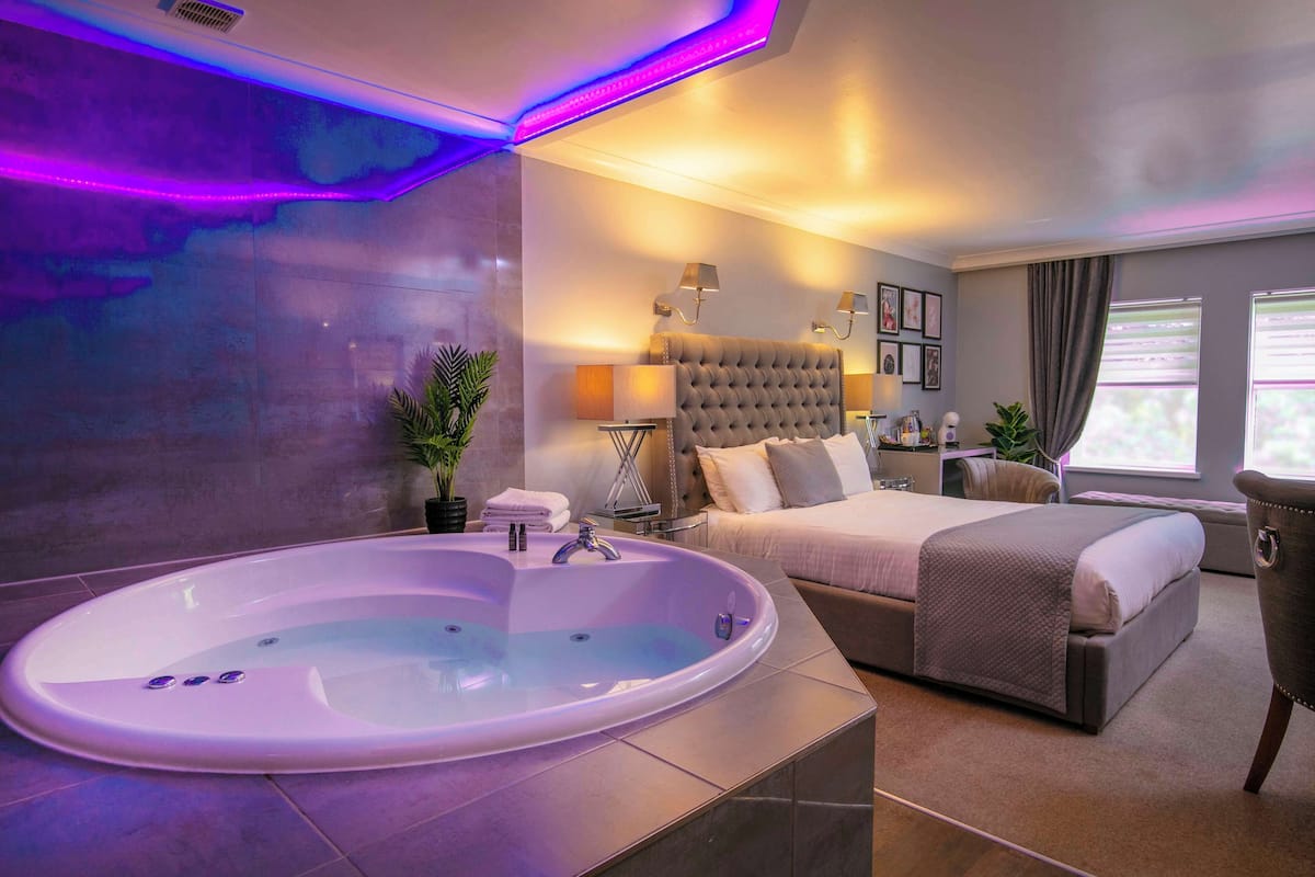 A modern hotel room with a large oval bathtub, a comfortable bed, a bedside table, and a view of the outdoors.