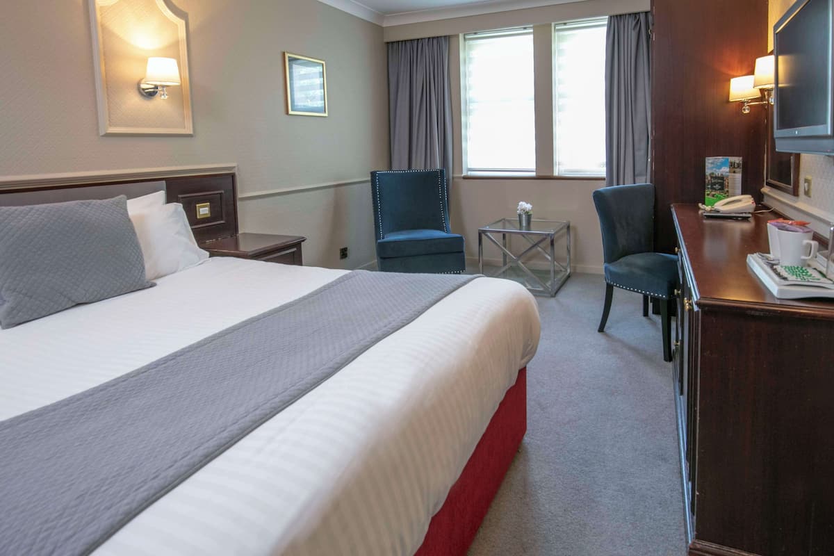 A hotel room with a large bed, two chairs, a small table, a desk with a telephone and a coffee cup, and a TV.
