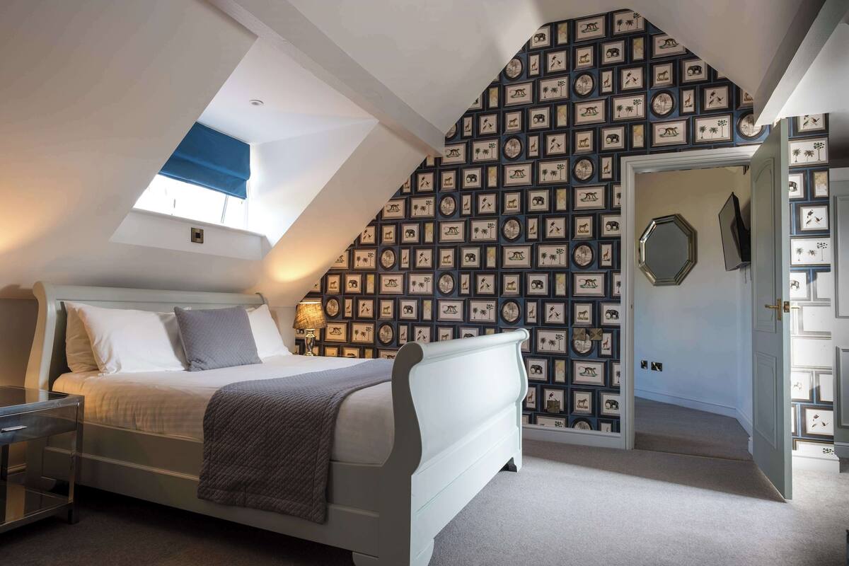 A bedroom with a sloped ceiling, a bed, a bedside table, a lamp, and a wall with framed pictures.