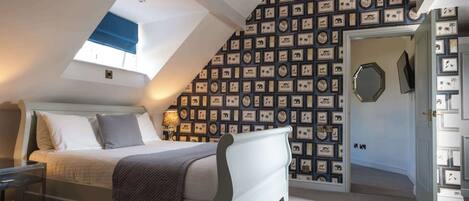 A bedroom with a sloped ceiling, a bed, a bedside table, a lamp, and a wall with framed pictures.