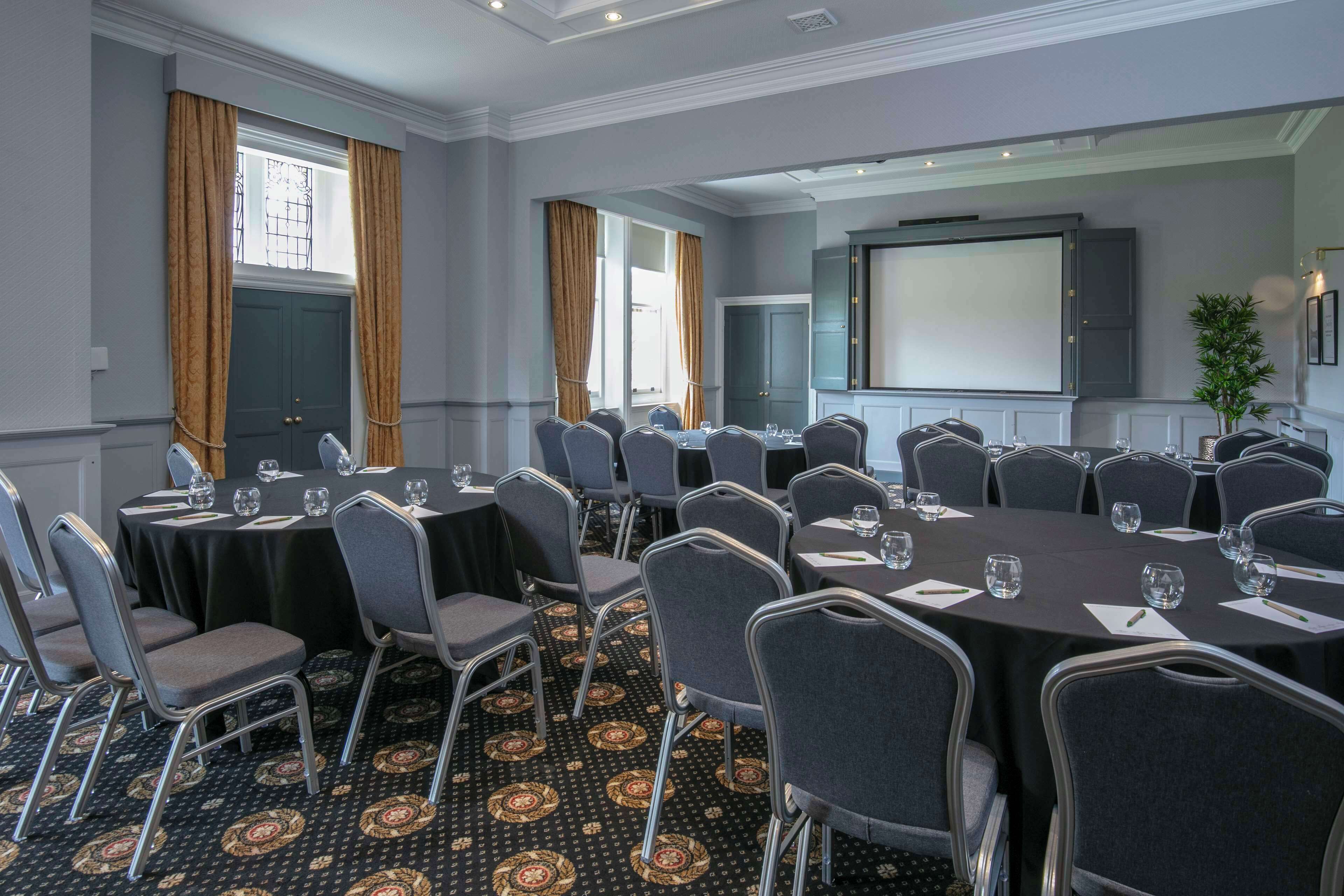 A conference room with multiple tables and chairs, a projector screen, and a large window with curtains.