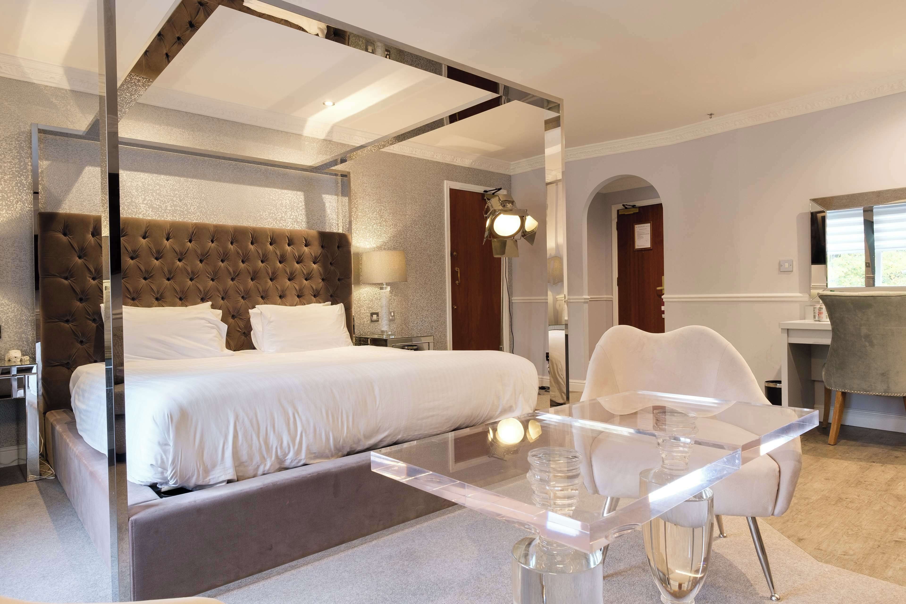 A modern hotel room with a large bed, a glass dining table, and a separate seating area.