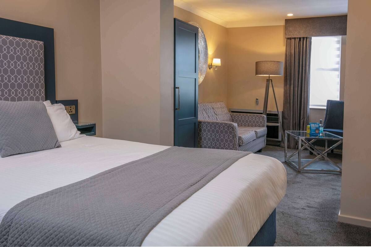 A hotel room with a large bed, a desk with a chair, a lamp, and a view of the outside through a window.