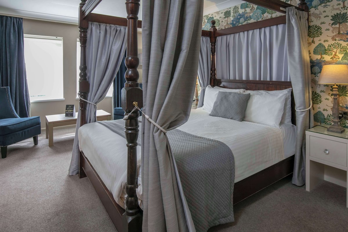 A four-poster bed with a canopy, a bedside table with a lamp, and a chair in a hotel room with floral wallpaper.