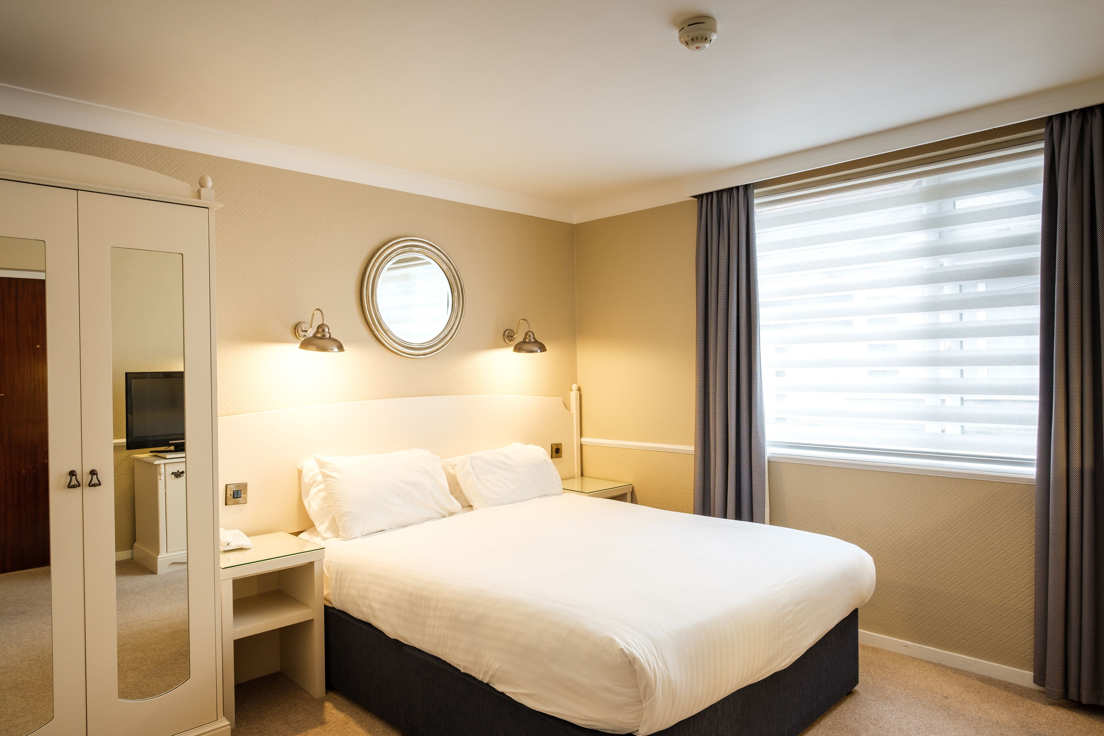 A hotel room with a bed, a desk, a mirror, a television, and a window with curtains.