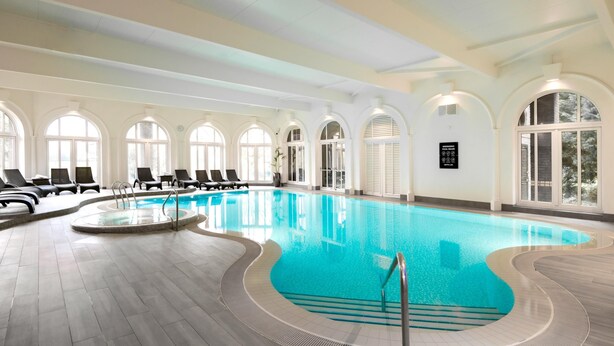 A large indoor swimming pool with a tiled floor, sunlit arched windows, and lounge chairs along the poolside. - Moor Hall Hotel, BW Premier Collection (Sutton Coldfield)