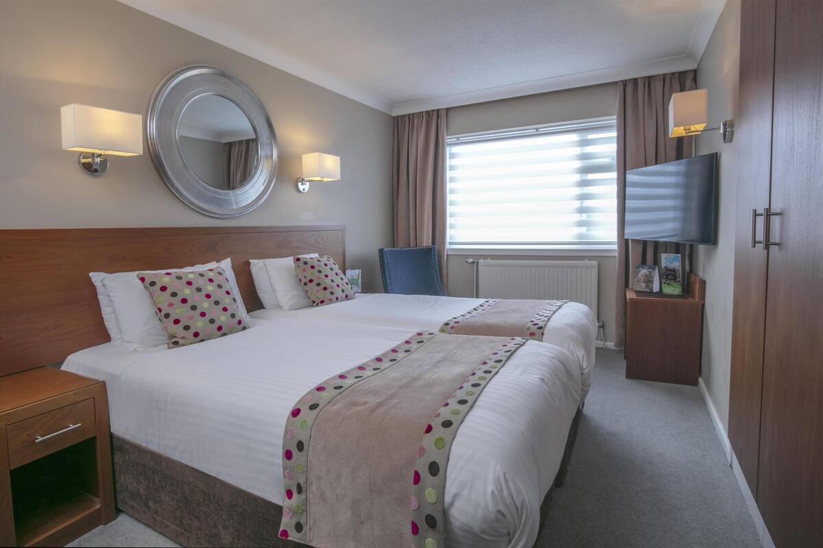 A hotel room with two beds, a large mirror, wall-mounted TV, and a window with blinds.