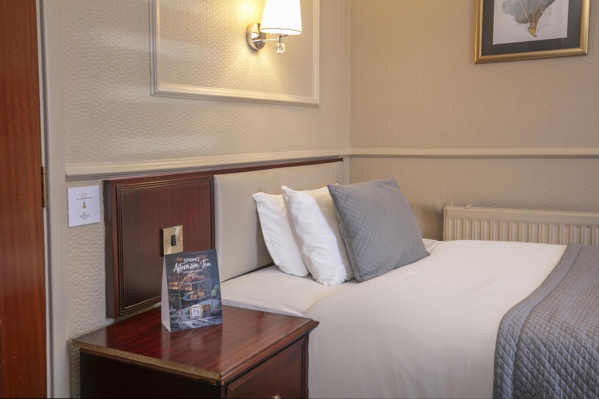 A hotel room with a neatly made bed, a nightstand with a framed picture, a wall-mounted lamp, and a framed artwork above the bed.