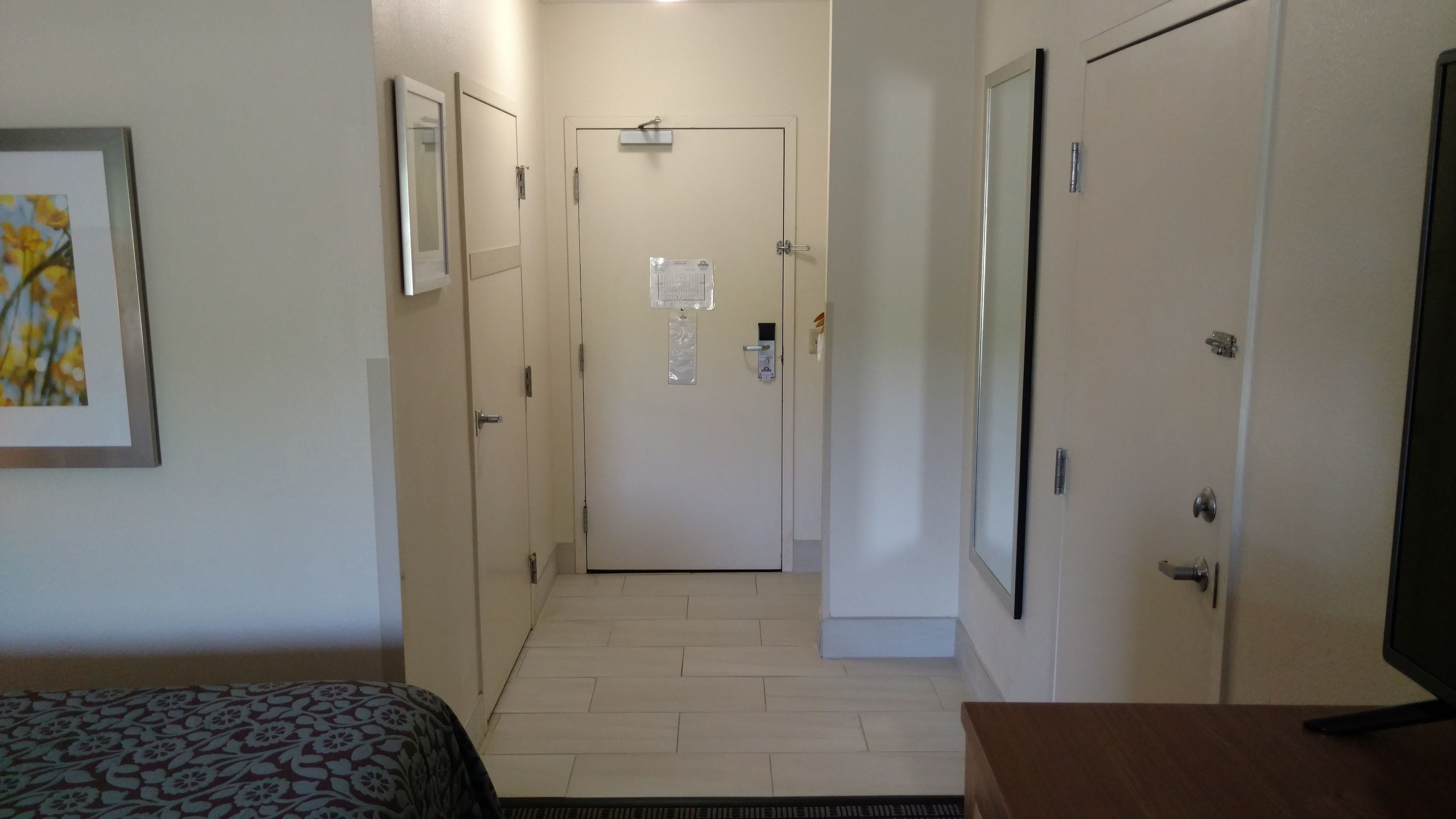 A narrow hallway with a bed on the left, a door with a security card reader in the center, and a TV on the right.
