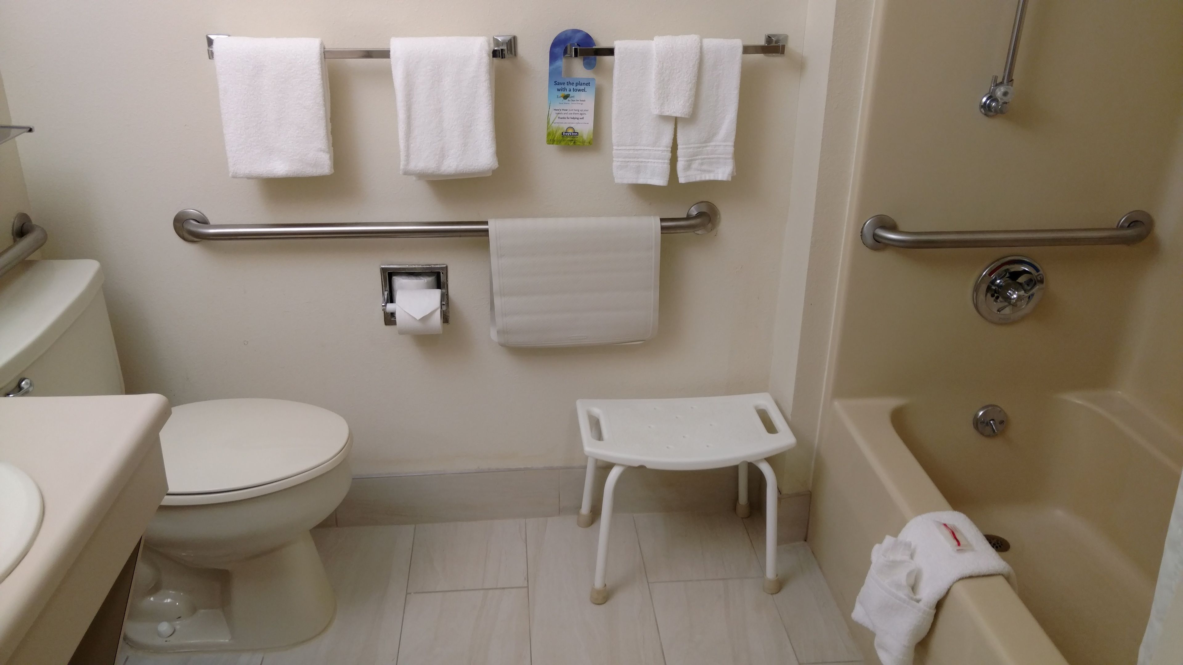 Un baño con inodoro, lavamanos, ducha y barra de apoyo. Varias toallas cuelgan de la pared.