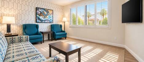 A living room with a patterned wall, two blue armchairs, a sofa, a coffee table, and a TV mounted on the wall.