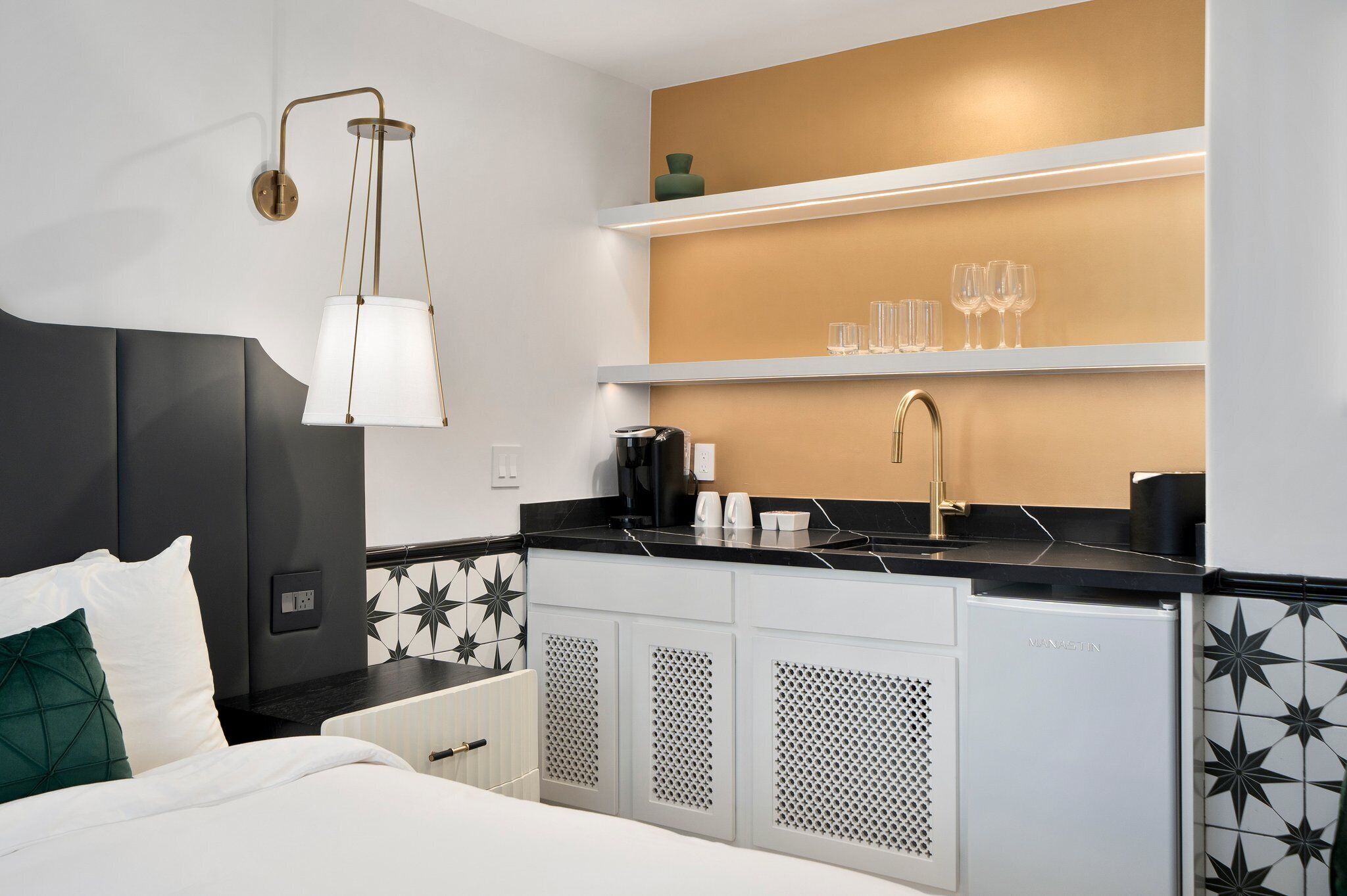 A modern hotel room with a bed, a minibar, a coffee machine, and a shelf with glasses.